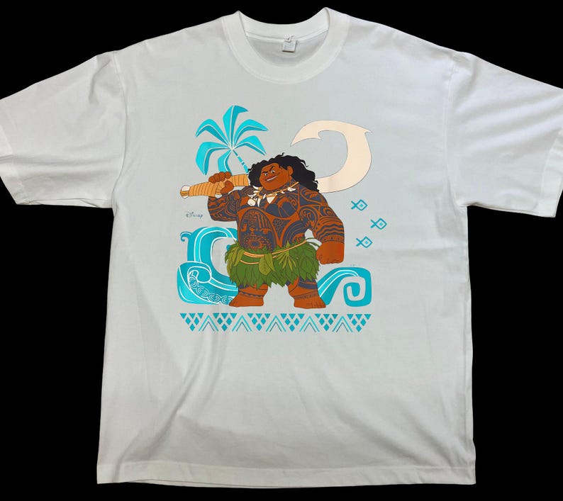 Puede incluir: Camiseta blanca con una ilustraci&oacute;n de dibujos animados de un personaje polinesio sosteniendo un arma. El personaje tiene piel morena, tatuajes y una falda de hierba. El dise&ntilde;o incluye una palmera, olas y peces en tonos azules y turquesas.