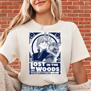 Puede incluir: Camiseta color crema con un gr&aacute;fico azul marino de Kristoff y Sven de Frozen II, con el texto "LOST IN THE WOODS". El dise&ntilde;o est&aacute; enmarcado por elementos decorativos y los nombres Kristoff Bjorgman & Sven Frozen II.