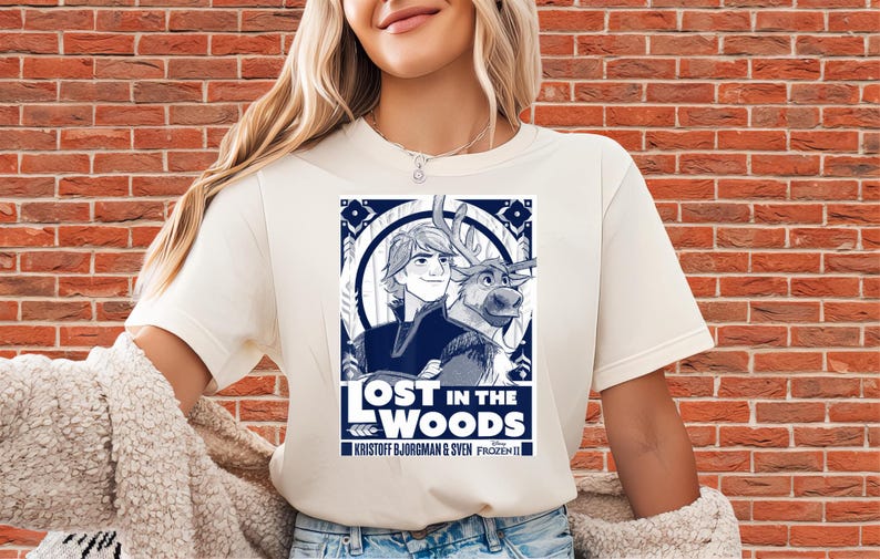 Puede incluir: Camiseta color crema con un gr&aacute;fico azul marino de Kristoff y Sven de Frozen II, con el texto "LOST IN THE WOODS". El dise&ntilde;o est&aacute; enmarcado por elementos decorativos y los nombres Kristoff Bjorgman & Sven Frozen II.