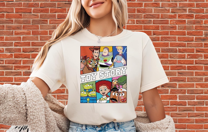 May include: Cream-colored t-shirt featuring a colorful graphic of characters from the animated film "Toy Story." The design includes Woody, Buzz Lightyear, and other characters, with the words "TOY STORY" in bold letters.