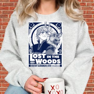 Puede incluir: Una sudadera gris claro con un dise&ntilde;o gr&aacute;fico azul y blanco de la pel&iacute;cula Frozen II. El dise&ntilde;o gr&aacute;fico incluye el texto "LOST IN THE WOODS" e im&aacute;genes de Kristoff, Sven y otros elementos de dise&ntilde;o. Una taza blanca con "XO XOXO" se sostiene en la mano.