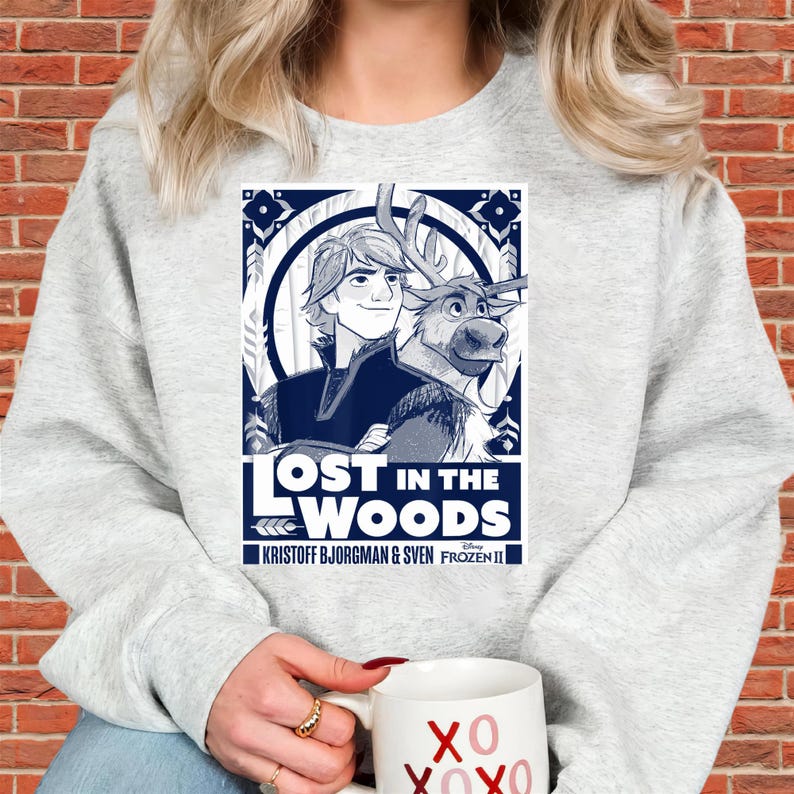 Puede incluir: Una sudadera gris claro con un dise&ntilde;o gr&aacute;fico azul y blanco de la pel&iacute;cula Frozen II. El dise&ntilde;o gr&aacute;fico incluye el texto "LOST IN THE WOODS" e im&aacute;genes de Kristoff, Sven y otros elementos de dise&ntilde;o. Una taza blanca con "XO XOXO" se sostiene en la mano.