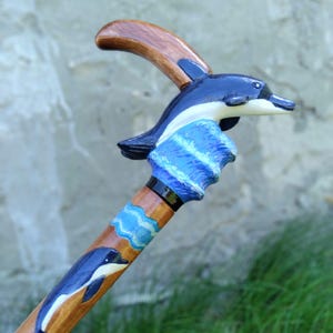 Puede incluir: Un bastón de madera con un tema de delfines. El mango curvo es marrón. La parte superior del bastón presenta un delfín tallado en negro, blanco y azul, emergiendo de olas azules. El eje es marrón con delfines y nubes pintados.