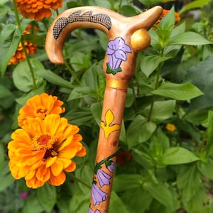 Pode incluir: Uma bengala de madeira com um cabo curvo apresentando um padrão texturizado de linhas cruzadas, um detalhe de esfera dourada lisa e um design pintado de flores roxas em forma de sino e um motivo de flor-de-lis amarela na haste.
