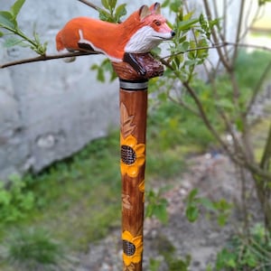 May include: A wooden walking stick with a carved fox head and a brown shaft decorated with sunflowers. The fox is orange and white, perched atop the handle. The shaft is brown with yellow sunflowers and leaf carvings.