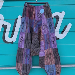 May include: Patchwork trousers in shades of purple, brown, and blue hang on a hanger. The trousers feature a mix of striped and solid fabric panels with floral designs. They have a drawstring waist and a relaxed fit.