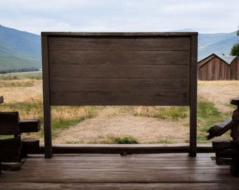 Puede incluir: Un letrero de madera con un acabado rústico, montado en un marco de madera. El letrero está en blanco y tiene un aspecto desgastado. El letrero está situado sobre un fondo de campo de hierba y montañas en la distancia.