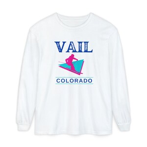 May include: White long-sleeve shirt with a graphic design featuring the word "VAIL" in blue, a skier in pink, and the word "COLORADO" in blue. The design is a retro ski resort theme.