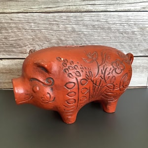May include: A terracotta-colored piggy bank with floral and leaf carvings. The bank has a slot on top for inserting coins and a smiling face. The piggy bank is a vintage item, with a rustic, aged appearance.