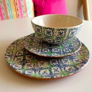 Puede incluir: Un cuenco de cerámica y dos platos con un estampado floral azul y verde. El cuenco y los platos están apilados. Los platos tienen un borde marrón. El fondo incluye cojines coloridos.