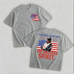 a grey t-shirt with a graphic design on the back, depicting a person wearing sunglasses and a racing helmet. The design includes an American flag and fireworks, along with the text "Raise Hell, Praise Dale" and "The American Spirit."