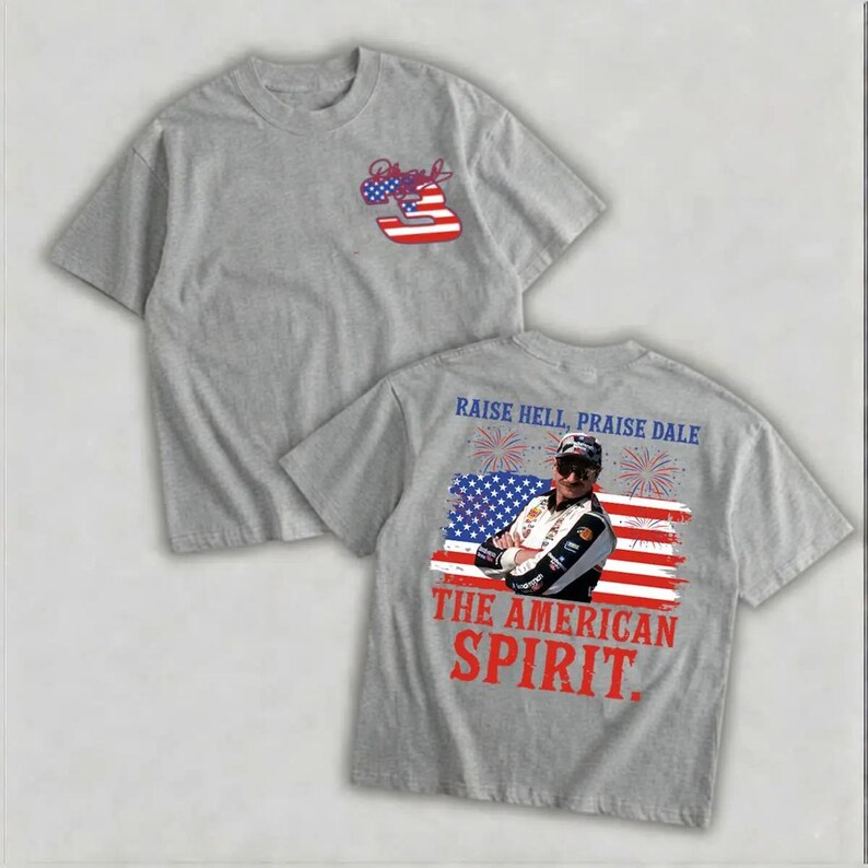 a grey t-shirt with a graphic design on the back, depicting a person wearing sunglasses and a racing helmet. The design includes an American flag and fireworks, along with the text "Raise Hell, Praise Dale" and "The American Spirit."