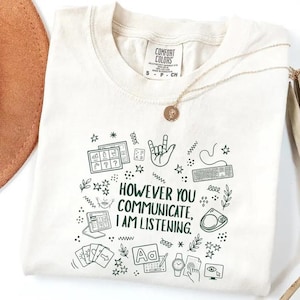 May include: Cream-colored t-shirt with the text "HOWEVER YOU COMMUNICATE, I AM LISTENING." The shirt features various hand-drawn icons, including a keyboard, hand sign, and cards. A gold necklace with a pendant rests on the shirt.