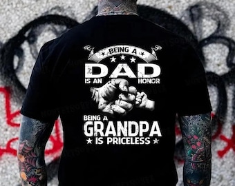 May include: A black t-shirt featuring white text and a graphic of a large fist bumping a small hand. The text reads: BEING A DAD IS AN HONOR, BEING A GRANDPA IS PRICELESS. The design is centered on the back of the shirt, which has short sleeves.