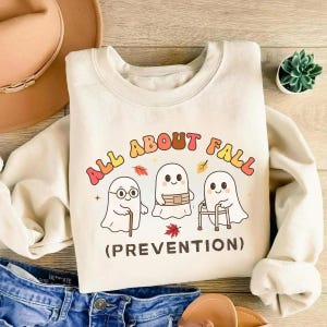 May include: A cream-colored crewneck sweatshirt featuring a graphic of three ghosts with the text ALL ABOUT FALL (PREVENTION). One ghost uses a cane, one wears a belt, and one uses a walker. The design includes small orange and red autumn leaf accents.