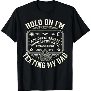 May include: A black short-sleeve t-shirt featuring a cream-colored graphic of a spirit board with the alphabet, numbers, and symbols. The text reads HOLD ON I'M TEXTING MY DAD, with a planchette icon centered at the bottom of the board design.