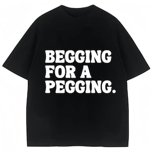 May include: Black t-shirt with the phrase "BEGGING FOR A PEGGING" in bold, white letters. The shirt has short sleeves and a crew neck. The text is centered on the front of the shirt.