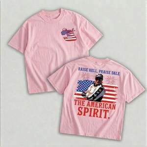 A pink t-shirt with a graphic design featuring an American flag and a person wearing a helmet, along with the text "Raise Hell, Praise Dale" and "The American Spirit."