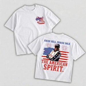 May include: White t-shirts with patriotic designs. One shirt features a stylized number 3 in red, white, and blue. The other shirt displays an image of a man in front of an American flag with the text "RAISE HELL, PRAISE DALE" and "THE AMERICAN SPIRIT."