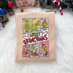 May include: A beige sweatshirt with a comic book style graphic of the Grinch. The graphic features the Grinch in different scenes with the text "Merry Grinchmas" and times like "4:00", "4:30", "5:00", "5:30", "6:30", and "7:00".