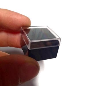May include: A small, square, black box with a clear plastic lid. The box is about 2 inches wide and 2 inches tall.
