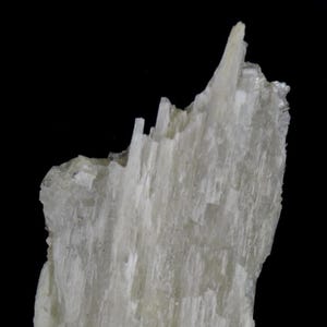 May include: A raw, off-white mineral specimen featuring a jagged, fibrous, and crystalline structure. The surface displays vertical striations and sharp, pointed edges that taper toward the top, set against a solid, plain black background.