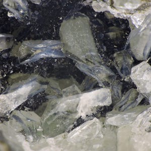 May include: A mineral specimen featuring translucent, pale blue to clear crystalline shards embedded within a dark, granular, charcoal-colored matrix. The jagged, angular crystal formations exhibit a glassy luster and varying degrees of transparency.