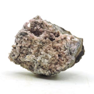 Pode incluir: Um espécime mineral bruto apresentando um aglomerado de cristais alongados em forma de lâmina, de cor rosa pálido, incrustados numa matriz rochosa rugosa de cor acastanhada acinzentada. A estrutura cristalina forma uma massa densa e texturizada.