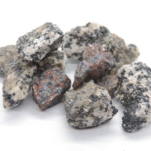 May include: A collection of rough-cut rocks with a mix of colors, including gray, white, and reddish-brown. The rocks have a speckled appearance with small, dark inclusions, and are displayed against a white background.