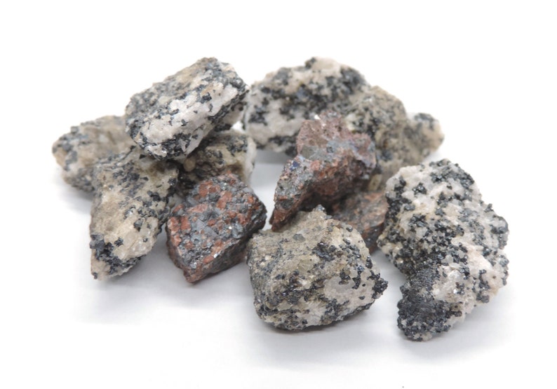 May include: A collection of rough-cut rocks with a mix of colors, including gray, white, and reddish-brown. The rocks have a speckled appearance with small, dark inclusions, and are displayed against a white background.