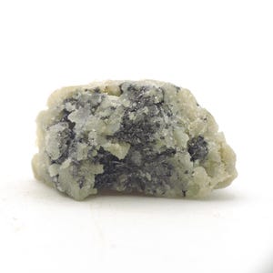 Könnte beinhalten: Ein unregelmäßig geformtes Mineralexemplar. Der Stein hat eine hellgrüne und cremefarbene Grundfarbe mit dunkelgrauen und schwarzen Einschlüssen. Die Oberfläche erscheint strukturiert und leicht kristallin.