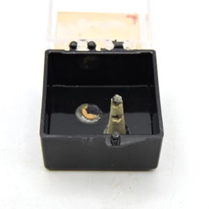 May include: A small black square plastic case with a hinged clear lid, containing a single off-white conical object with a dark tip and a small circular orange and grey disc-shaped component resting on the interior base of the container.
