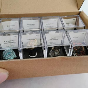 May include: A cardboard box containing eight clear plastic display cubes, each housing a unique mineral specimen. Each cube features a white label with printed text identifying the mineral, such as Carbonate-cyanotrichite, Stilbite, and Zincite.