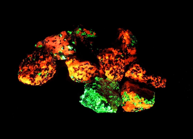 May include: A collection of glowing rocks against a black background. The rocks display vibrant orange and green colors, with the orange areas speckled with dark spots. The overall effect is a striking contrast of light and shadow.