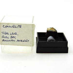 May include: A small mineral specimen of connellite displayed in a black plastic box. The specimen is dark with blue-green highlights. A clear plastic lid is attached to the side, featuring a white label with the text: CONNELLITE, TRIAL LEVEL, BULL BAY, AMLWCH, ANGLESEY.