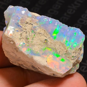 May include: A rough, natural opal with a milky white base and flashes of iridescent green, blue, and orange. The opal is still attached to its matrix.