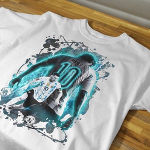 May include: White t-shirt featuring a blue and black graphic print of a football player wearing a jersey with the number "10" and the name "Messi" on it.