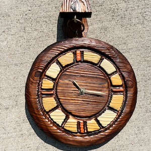 May include: A round wooden wall clock with a brown wood face and yellow and orange accents. The clock has a unique design with a carved wood frame and a hanging bracket.