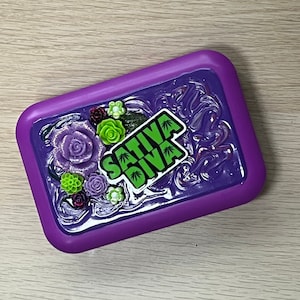 May include: Purple silicone rolling tray with a green and black "SATIVA DIVA" sticker and green and purple flower embellishments.
