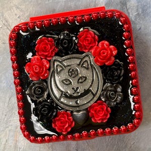 May include: A red and black decorative box with a cat design. The box is adorned with red and black roses and a silver cat emblem. The edges are lined with red beads, creating a gothic or alternative aesthetic.