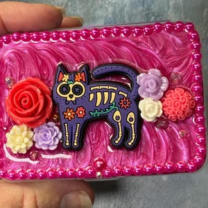 May include: A decorative trinket box with a vibrant pink background and a border of small pink pearls. The box features a purple cat skeleton design with floral accents and is adorned with colorful resin flowers.