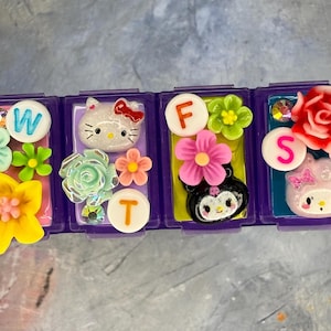 May include: A purple plastic pill organizer with four compartments decorated with colorful resin flowers, rhinestones, and character charms. The lids feature white circular labels with the letters W, F, S, and T, alongside miniature figures.