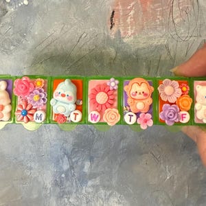 Puede incluir: Un pastillero semanal verde con siete compartimentos, cada uno decorado con un dise&ntilde;o diferente de animal y flor. Los d&iacute;as de la semana est&aacute;n etiquetados con letras en peque&ntilde;os c&iacute;rculos blancos.