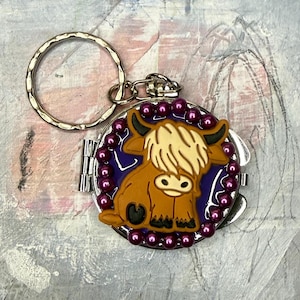 May include: A round, silver-colored keychain with a decorative charm. The charm features a cartoon Highland cow in shades of brown, tan, and black, set against a purple background and surrounded by small, purple beads. The keychain has a split ring.