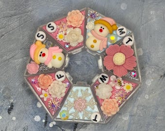 Op de afbeelding: Een doorzichtige pillenorganizer met meerdere compartimenten en een roze en glinsterend interieur. Elk compartiment is versierd met bloemen, sneeuwpoppen en de dagen van de week. De organizer heeft een unieke, geometrische vorm.