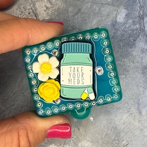 May include: A rectangular teal resin badge reel featuring a central illustration of a medicine bottle with the text TAKE YOUR MEDS, accompanied by a white flower and a yellow flower, all bordered by a frame of small clear beads and rhinestones.