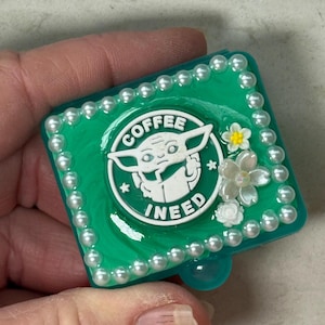 May include: A green square-shaped box with a white pearl border. The box has a green and white coffee cup design with a cartoon character inside. The text "COFFEE I NEED" is on the cup. The box is decorated with white and yellow flowers.