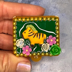 May include: A decorative, rectangular trinket box with a green background and a yellow zebra figure. The box is adorned with gold-colored beads and colorful flowers, including pink, white, and green roses. The zebra has black stripes.