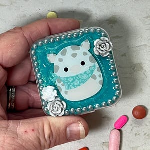 May include: A small turquoise blue pill box with a white and gray cartoon animal wearing a turquoise bandana. The box is decorated with silver and white flowers and silver beads.