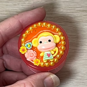May include: A round, orange and red plastic container with a gold beaded trim. The container features a 3D image of a monkey with a pink face, green and white striped shirt, and a red heart. The monkey is surrounded by pink, yellow, and blue flowers.
